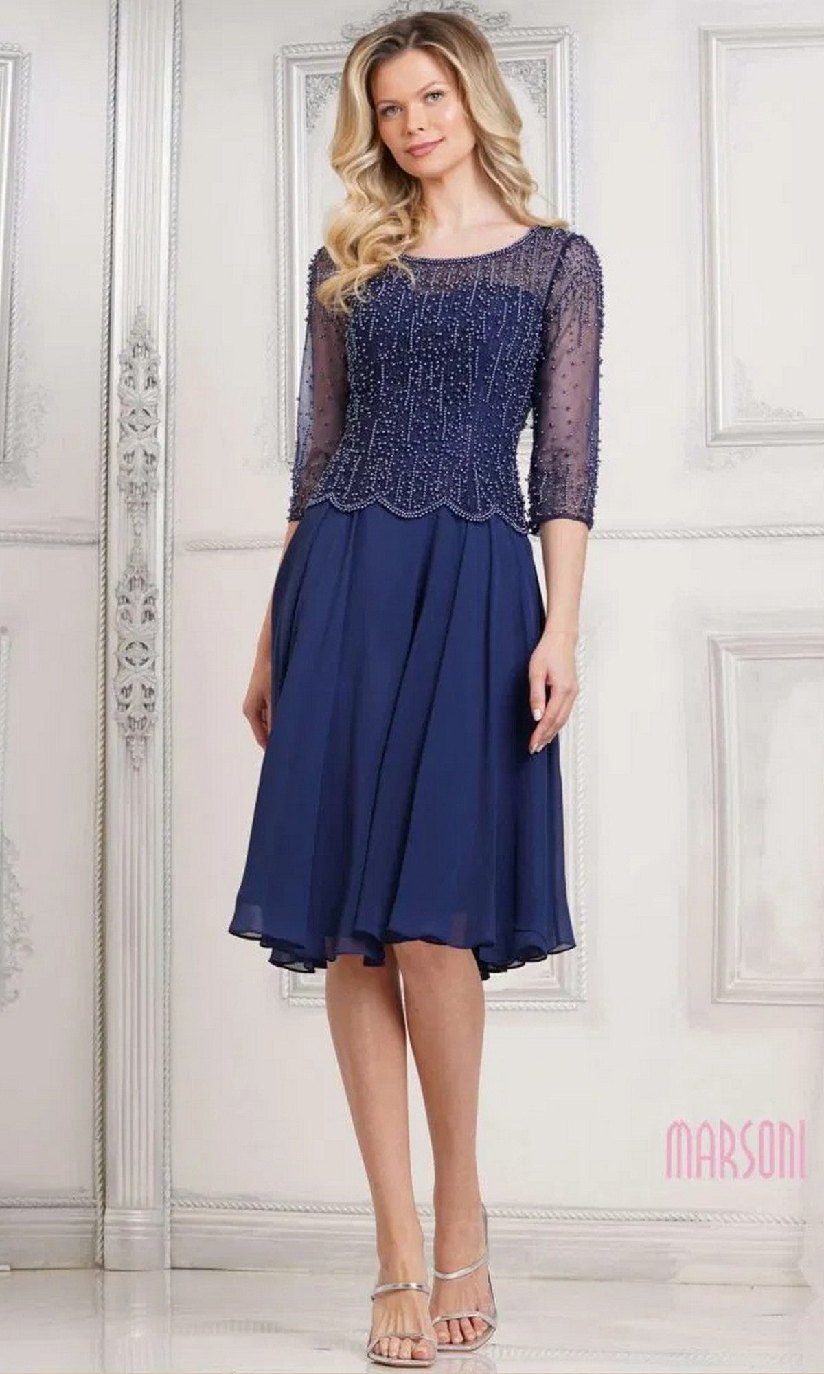 A woman wearing the Marsoni M312S knee-length semi-formal dress in navy, featuring a fit-and-flare silhouette, sheer beaded bodice, and three-quarter sleeves, stands against an ornate white wall.